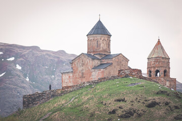 Fototapeta na wymiar Gergeti trinity church on the hill with priest standing in black clothes nearby and enjoying panorama. Religion and saint places in Georgia. 2020