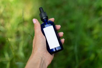 Fototapeta premium Against the background of green grass, a female hand holds a blue cosmetic bottle