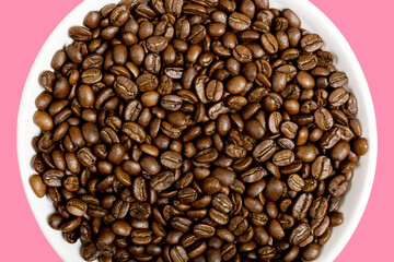 Fragrant coffee beans on a white plate on a pink background. Beautiful background and texture. Coffee shop or store concept. Top view, closeup.