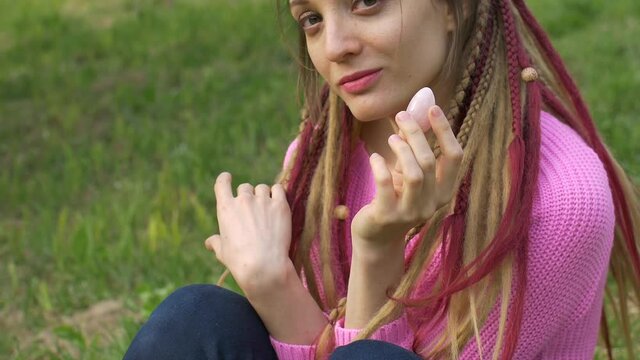 Adult girl in knitted sweater and long dreadlocks is holding pink quartz yoni egg for vumfit, imbuilding or meditation outdoors on her body background outdoors