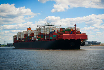 A cargo ship with many colorful containers floats along the river on a sunny day.