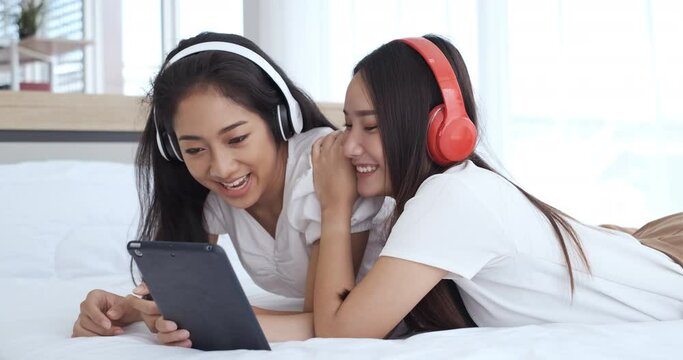Two young asian women lying in bed and wearing headphones doing video call