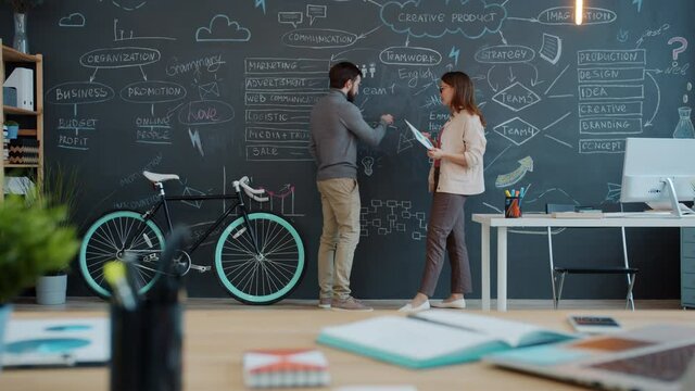 Girl and guy are talking and writing on blackboard wall in shared office planning business strategy discussing work. Occupation and interior concept.