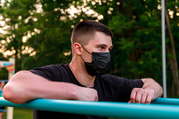 Sports during quarantine, self-isolation in urban conditions. Young guy, an athlete engaged on uneven bars. He's wearing black medical mask. Covid-19 coronavirus pandemic: life of active person