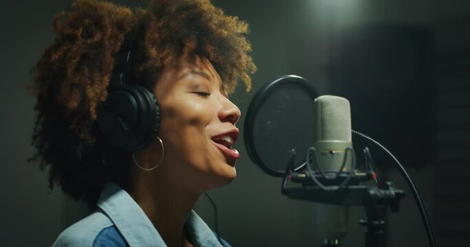 An young professional smiling energetic african female singer wearing headphones is performing a new song with a microphone while recording it in a music studio.