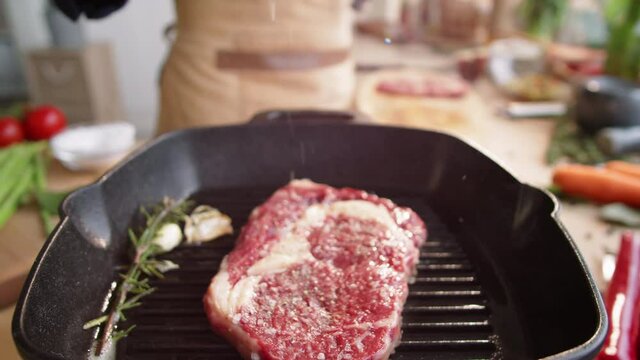 Zoom out shot of male chef pouring salt over meat steak on hot grill pan with rosemary and garlic cloves on it while cooking in the kitchen
