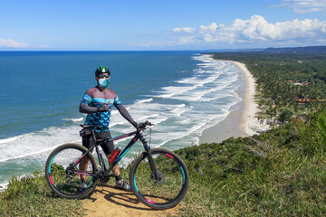 Coronavirus (COVID-19). Young man on MTB bike wearing protective mask doing pose alone in beautiful landscape with beaches in the background.