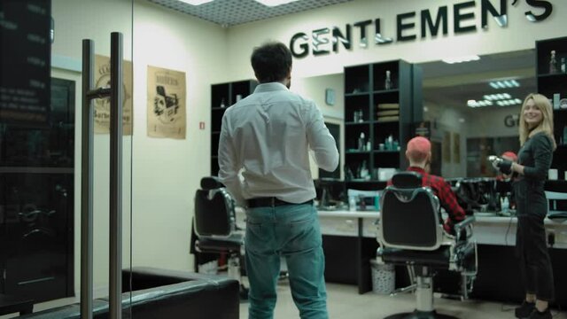 Tracking shot of bearded man smiling and greeting barbers then taking seat on comfortable leather sofa during visit in modern barbershop.