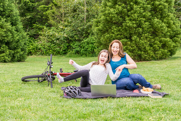 Beautiful pregnant woman and her daughter are laughing in a summer green park with laptop and bicycle.