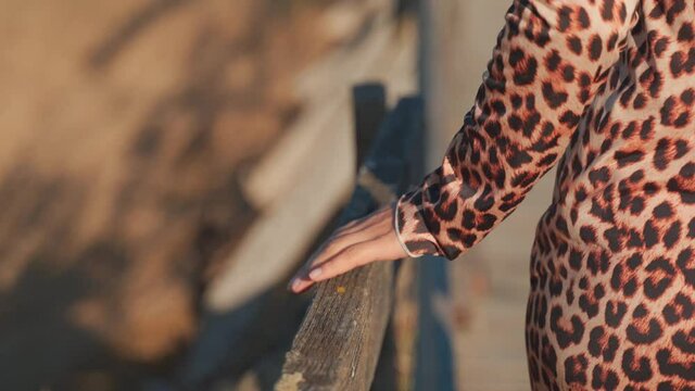 Caucasian woman wearing animal printed dress walking on pathway gliding hand across outdoor wooden handrail in countryside, handheld behind close up