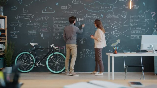 Man and woman coworkers are writing on chalk board discussing business strategy in open space office. Interior, coworking space and marketing concept.
