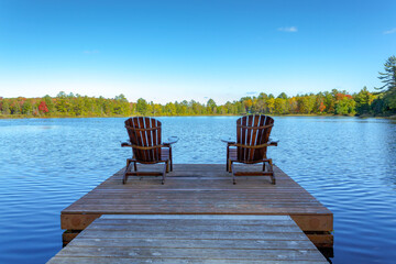 Two Muskoka chairs sitting on a wood dock facing a lake in a calm autumn season sunny day, with the...