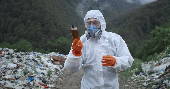 Man in protective suit walking near garbage pails in forest. Male person in respirator and gloves carrying and looking at plastic bottles. Concept of environmental pollution