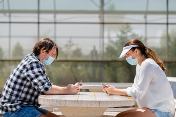 young couple wearing surgical masks and sitting at a table in front of each other using their phones