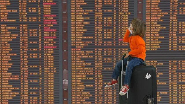 Funny little child with mask sit on big suitcase in front of departure airport, safe corona virus travel, blond hair boy with snickers waiting