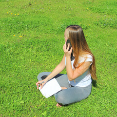 A beautiful young white girl in a white t-shirt and with long hair sitting on green grass, on the lawn and working behind a black laptop and talking on a cell phone and writes with a pen in a notebook