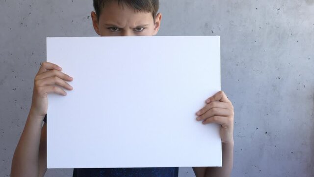 Boy holding in his hands blank empty white mock up board poster. Sad kid covering his face with canvas board. Grey wall on background