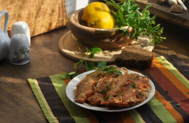 Fried fish on a plate on an old wooden table. Lemons and greens in a wooden bowl. The old village house interior. Wooden window.