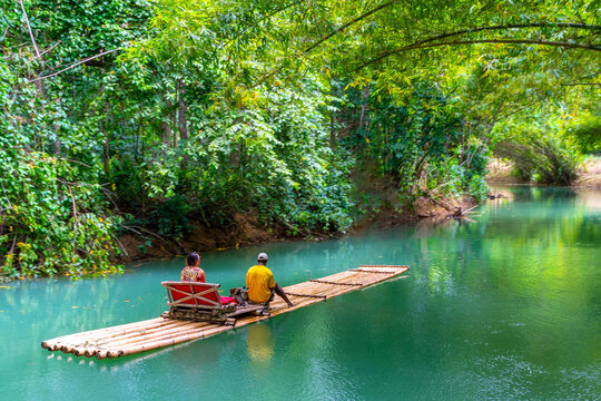 Female tourist and tour guide sit on bamboo raft on Martha Brae River in Falmouth, Jamaica. Relaxing scenic ride through quiet countryside landscape under canopy of trees. 