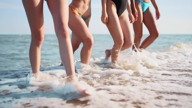 Slim female legs and feet walking out of sea water waves on sandy beach. Pretty women move on seafront. Splashes of water and foam in 120 fps slow motion. Girls after bathing in ocean go to the shore.
