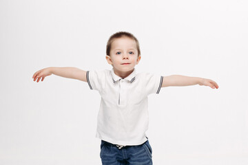 Funny little boy with blue eyes, wearing in white polo t-shirt, jeans, gesturing by hands and showing wings like flying bird, in different sides. Kid playing at studio and enjoying. White background.