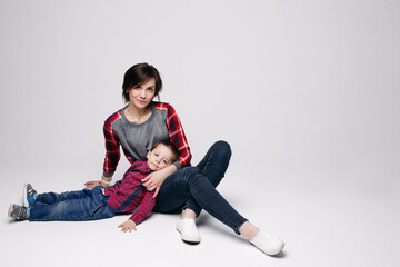 Obraz na płótnie Canvas Happy mother and little son in red checked shirt and jeans, spending time together, embracing, leaning each other. Family of mom carrying about child and smiling at camera. Gray studio background.