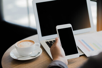 Phone in hand on table with coffee cup and laptop in office.