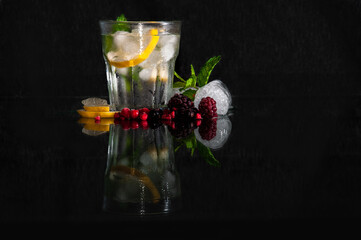 A glass with water, ice, a slice of lemon and a leaf of mint is on a glass table with reflection on a black background, next to a glass are red berries, slices of lemon, ice cubes.