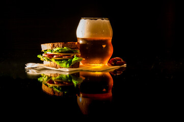 A glass of beer and a sandwich with cheese, sausage and salad on a napkin are on a glass table with reflection on a black background