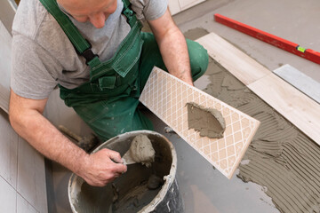 The builder applies cement adhesive to the ceramic tile with a stainless trowel.