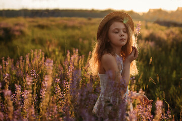 A baby girl in a straw hat stands in a field of flowers at sunset in summer