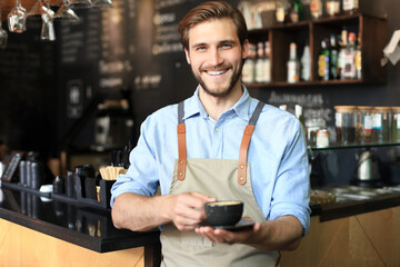 Handsome barista offering a cup of coffee to camera at the coffee shop. - obrazy, fototapety, plakaty
