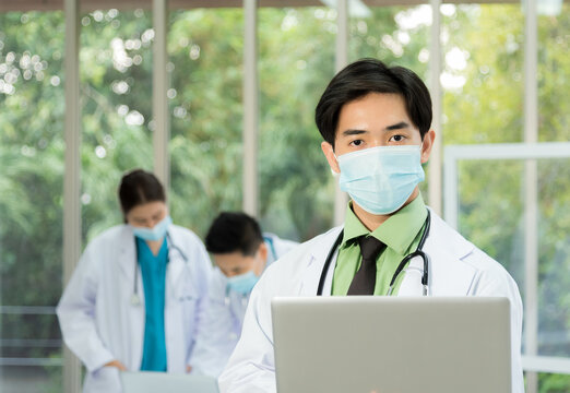 Profesional doctor wearing mask, lab coat and stethoscope standing in front of staff team and holding laptop in conference room