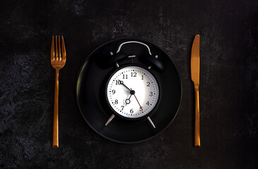 Alarm clock on a black plate with copper knife and fork. Intermittent fasting concept