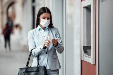 Fototapeta na wymiar Elegant woman with protective mask standing on city street and using alcohol spray to disinfect her hands after use of ATM machine. Corona or Covid-19 virus pandemic prevention and healthcare concept.