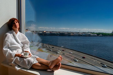 Middle-aged woman in white bathrobe enjoying sunny river view next to big window with cup of coffee. Panoramic window with blue sky and urban landscape with river.