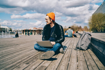 Positive male student spending time on publicity area with modern laptop computer using for e learning and communicate, happy hipster guy dressed in stylish outfit enjoying sunny autumn weather