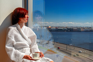 Middle aged woman in white bathrobe enjoying sunny river view next to big window with cup. Panoramic window with blue sky and urban landscape with river.
