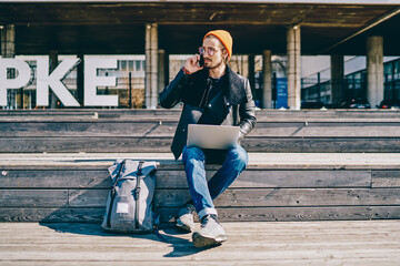 Serious hipster guy dressed in fashionable clothing making international call to operator for consultancy during distance job outdoors, millennial man sitting with netbook and talking via cellphone