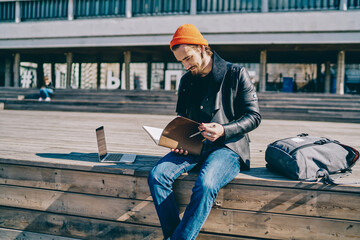 Millennial male student in stylish outfit reading information from education textbook spending time for e learning, trendy dressed intelligent hipster guy holding sketchbook for designing in hands