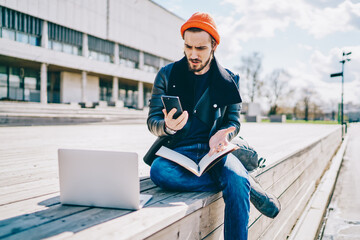 Confused Caucasian male student puzzled with received web information with exams schedule, millennial hipster guy dressed in trendy clothing reading message during e learning at urban setting