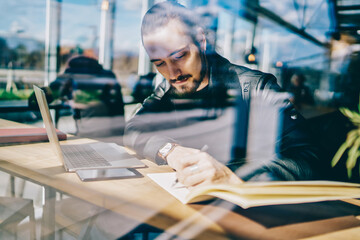 Creative male freelancer concentrated on making sketch in notebook sitting in cafe with laptop computer for online work, pensive hipster guy writing ideas for article publication in blog near window.