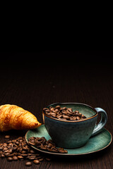 Cup with coffee beans on a black background. Freshly roasted arabica. Fresh croissant on a wooden table.