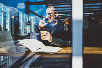 Cheerful handsome blogger working in cafe sitting near window talking on mobile phone, happy hipster guy calling friend invites for having cup of morning coffee and work online with laptop computer .