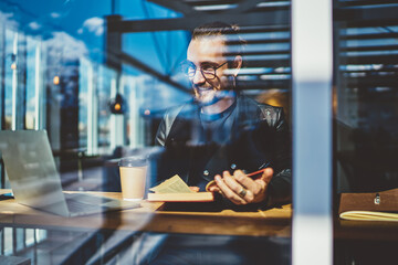 Cheerful handsome hipster guy in spectacles enjoying morning sitting in coffee shop with interesting book, happy prosperous male freelancer feeling carefree satisfied with remote job in cafe.