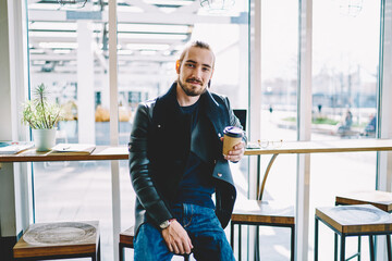 Portrait of Caucasian hipster guy holding paper cup with hot caffeine beverage and looking at camera during leisure time, millennial male teenager dressed in stylish apparel enjoying break for coffee