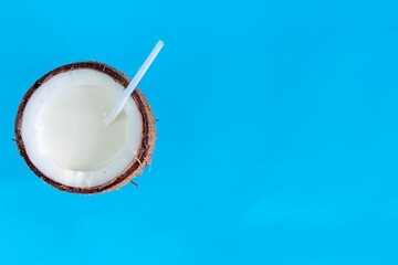 Brown half of coconut nut with white milk and straw inside on blue background. Tropical exotic fruit, cocktail, water for refreshment. Organic natural healthy vegan meal, coco based food concept.