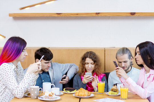 Young people guys and girls are having meeting, sitting at table in cafe. Friends are holding smartphones and browsing social networks instead of speaking. Addiction to gadgets, internet concept.