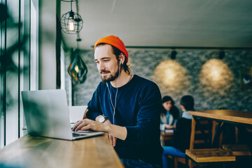 Caucasian male resting in coffee shop listening favorite music in headphones and chatting in social networks, skilled freelancer enjoying remote job typing publication for web blog on laptop