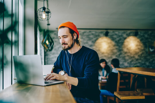 Caucasian Male Resting In Coffee Shop Listening Favorite Music In Headphones And Chatting In Social Networks, Skilled Freelancer Enjoying Remote Job Typing Publication For Web Blog On Laptop